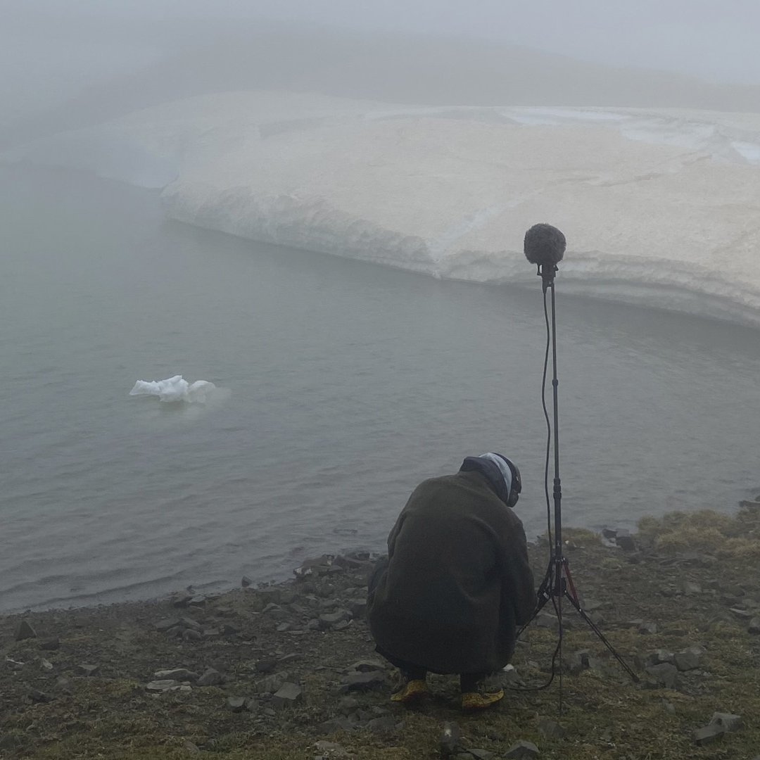 Stefanos Levidis in winter gear squatting next to a professional microphone on a high stand, on the shore of a snowy lake Gistova.